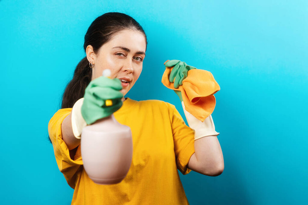 Portrait of a beautiful Caucasian young woman in rubber gloves, holding a rag in her hands and shoots of sprayer. Blue background. Concept of cleaning service. - Valokuva, kuva