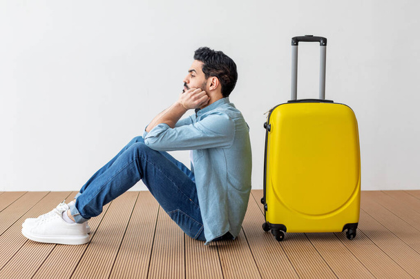 Upset arab male tourist sitting near suitcase and looking away at copy space, waiting for flight over light wall - 写真・画像