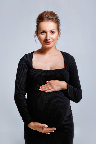 Portrait of a pregnant woman in a tight black dress on a gray background. The woman gently holds her stomach with her hands. - Foto, Imagem