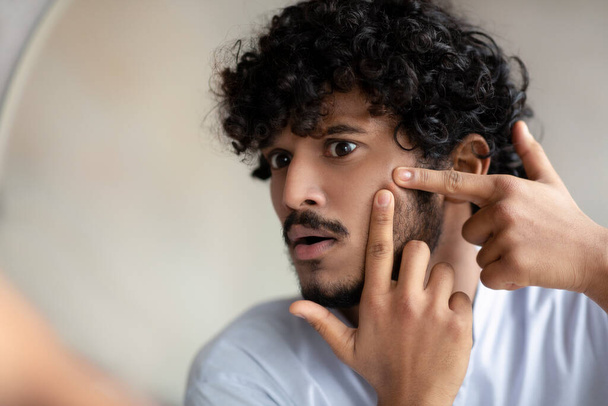 Egy jóképű indiai férfi arcképe, aki megérinti az arcát, ujjaival pattanást nyom az arcára, tükörbe néz, közelről - Fotó, kép