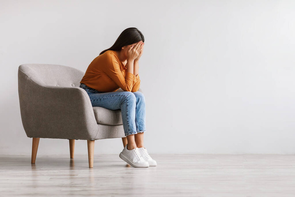 Jovem mulher asiática enterrando o rosto em mãos e chorando, sentando-se deprimida, sentada em poltrona contra a parede do estúdio branco - Foto, Imagem