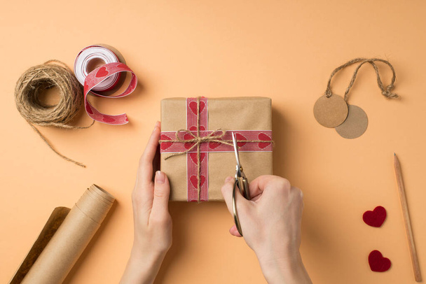 First person top view photo of valentine's day decor female hands cutting twine on craft paper giftbox with scissors roll of kraft paper spool of ribbon pencil tags hearts on isolated beige background - Photo, Image