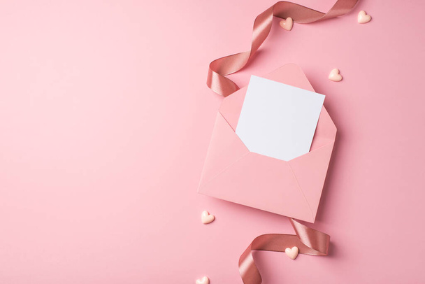 Вид сверху фото день святого Валентина украшения кудрявая шелковая лента открываются розовый конверт с бумажным листом и маленькие сердца на изолированном пастельно-розовом фоне с копирайтом - Фото, изображение