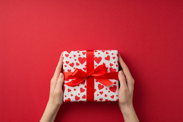 Première personne vue du dessus photo de la Saint-Valentin décor mains de jeune femme donnant boîte-cadeau en papier d'emballage blanc avec motif coeur et arc rouge sur fond rouge isolé avec espace vide - Photo, image