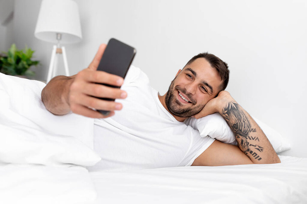Усміхаючись тисячолітній привабливий хлопець з бородою лежить на білому ліжку, спілкується і читає новини на смартфоні
 - Фото, зображення