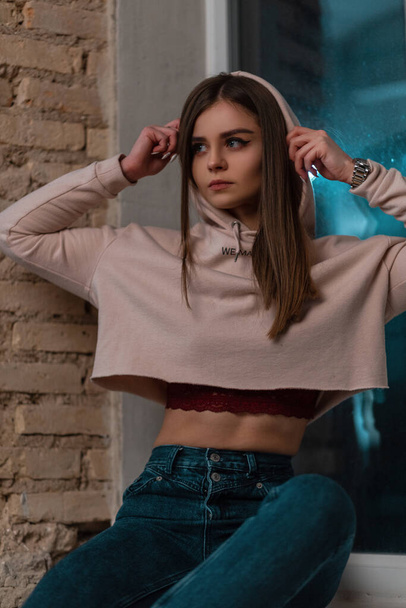 Stylish model of a young woman in fashionable clothes puts on a hood. Beautiful pretty trendy girl in a pink sweatshirt in jeans sits on a windowsill near a vintage brick wall. Cute lady. Casual style - Zdjęcie, obraz