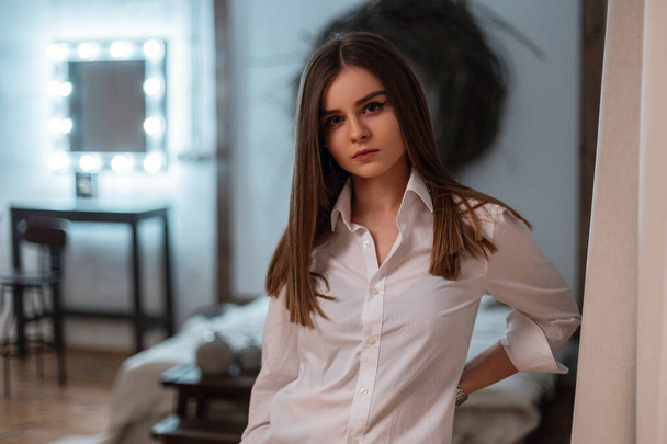 Pretty modern young woman fashion model in a classic stylish white shirt resting in the vintage cozy bedroom. Attractive trendy girl with brown hair in a elegant clothes is posing in the room. Style. - Фото, изображение