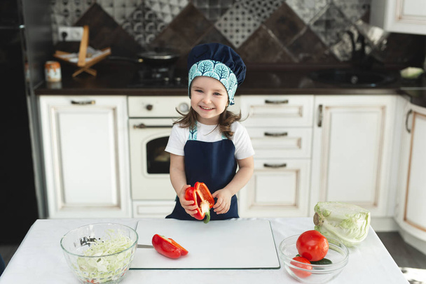 A child in an apron and a chefs hat in the kitchen is preparing a salad, eating red pepper and laughing - Foto, afbeelding