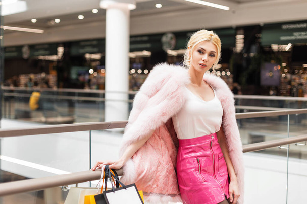 Moderne junge Frau in modisch eleganter Kleidung mit bunten Papiertüten, die sich nach dem Einkauf in der Nähe einer Glaswand in einem Geschäft ausruhen. Attraktive stilvolle sexy Mädchen Mode-Modell. - Foto, Bild