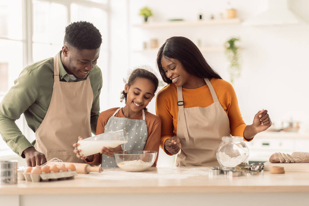 Musta tyttö lisäämällä maitoa taikina leipominen vanhempien kanssa sisätiloissa - Valokuva, kuva