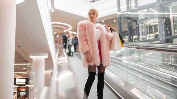 Modern young woman with blond hair in fashionable elegant clothes in black shoes with multi-colored paper bags rides on an escalator in store. Attractive stylish girl fashion model in luxury outfit. - Photo, Image