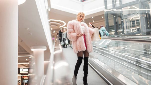 Stylish cute young blonde woman in fashionable luxurious fur coat in elegant vintage black shoes with shopping bags poses on escalator in modern store. Sexy girl in expensive clothes in shop mall. - Фото, зображення