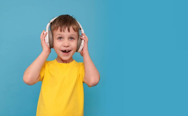Happy child in a yellow t-shirt on a blue background with headphones on his head - Photo, Image