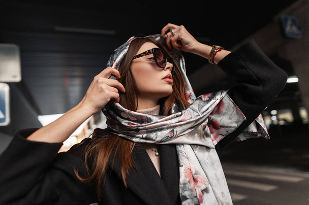 Κομψό νεαρή όμορφη γυναίκα σε κομψό σκούρα γυαλιά ηλίου σε μαύρο παλτό βάζει ένα πολυτελές μεταξωτό μαντήλι στο κεφάλι σε εξωτερικούς χώρους. Όμορφο μοντέλο μόδας κορίτσι χαλαρώνει στο δρόμο στην πόλη. Σέξι κυρία.. - Φωτογραφία, εικόνα