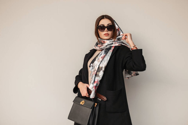 Gorgeous pretty young professional woman in stylish sunglasses in trendy black coat with leather bag with a vintage scarf on head posing near a wall outdoors. Attractive girl fashion model. Sexy ladie - Photo, Image