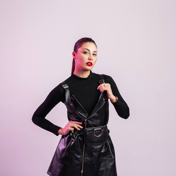 Gorgeous young brunette woman with trendy hairstyle in vintage shirt in leather fashionable black sexy dress posing in the studio. Attractive girl with beautiful red lips indoors. Clothes for women. - Fotó, kép