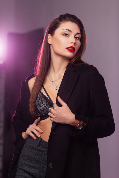 Гламурний портрет молодої привабливої жінки з пухнастими сексуальними червоними губами з довгим коричневим волоссям в модному мереживному бюстгальтері в стильному чорному пальто в студії з яскравим світлом.
 - Фото, зображення