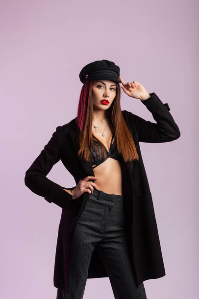 Modern lány divat modell egyenesíti divatos fekete sapka beltérben. Stílusos fiatal nő vintage kabát elegáns csipke fehérnemű divatos nadrág szexi piros ajkak a stúdióban a fal közelében. - Fotó, kép