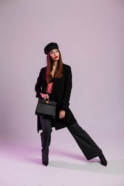 Модна модель молодої жінки в елегантному мереживному бюстгальтері в куртці в штанях в чоботях зі шкіряною сумкою стоїть в кімнаті біля стіни. Сексуальна дівчина з коричневим волоссям позує в стильному чорному одязі в приміщенні
 - Фото, зображення