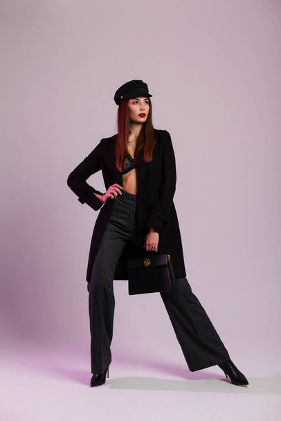 Business luxusní mladá žena s červenými sexy rty v krajkové podprsenky ve stylové čepici v dlouhé bundě v pruhovaných kalhotách v elegantních botách s koženou módní kabelkou pózuje ve studiu. Atraktivní dívčí model. - Fotografie, Obrázek