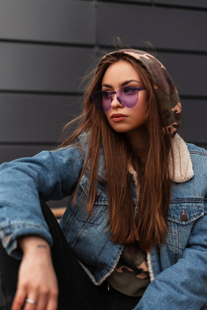Modern American jeune femme hipster dans des lunettes violettes élégantes dans la jeunesse veste en denim bleu se détendre à l'extérieur près de bâtiment gris. Belle fille modèle en tenue décontractée dans la rue. Printemps style jeunesse. - Photo, image
