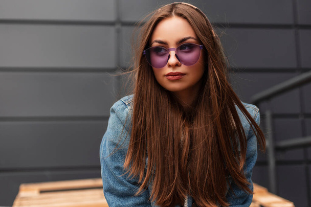 Retrato elegante jovem legal com lábios sexy hipster em óculos violeta em casaco de ganga vintage azul elegante perto do edifício cinza ao ar livre. Modelo menina urbana está descansando na cidade. Moda de rua - Foto, Imagem