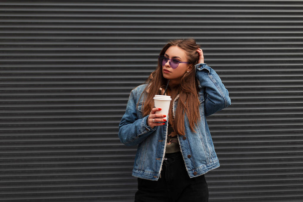 Modern modern hippi kadın şık gençlik kotları ve modaya uygun mor gözlükleriyle kentin metal bir duvarının yanında duran bir fincan kahveyle. Amerikalı kız model açık havada. - Fotoğraf, Görsel