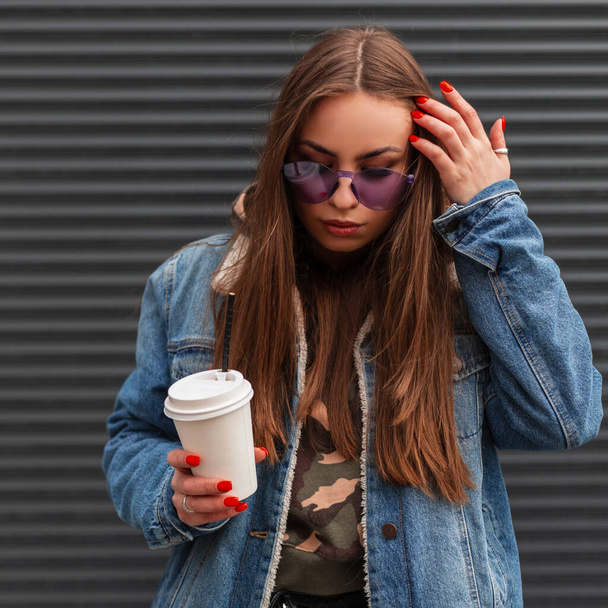 American young hipster woman in stylish glamorous purple glasses in trendy blue denim jacket with coffee straightens hair and looking down near vintage metal wall. Cute girl fashion model outdoors. - Foto, Imagen