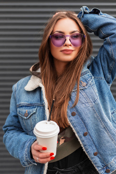 Modelo jovem feliz atraente em óculos roxos na moda em casaco de ganga elegante com xícara de bebida quente posando perto de uma parede de metal ao ar livre. Menina hipster bonita com café está desfrutando de dia de primavera. - Foto, Imagem