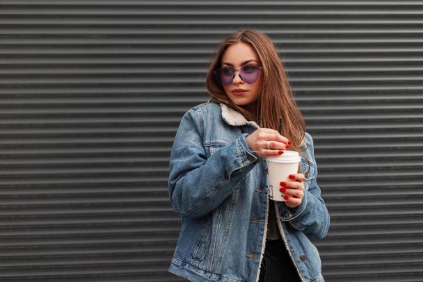 Europeu bonito jovem hipster mulher na moda jaqueta azul jeans em óculos roxos da moda com xícara de café quente está de pé perto de uma parede cinza na cidade. Menina bonita posando na rua. - Foto, Imagem