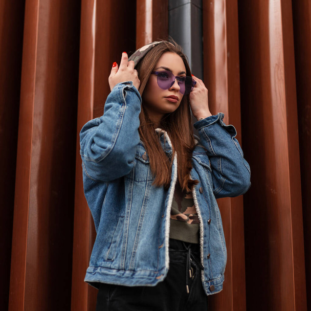 Modna młoda hipsterka zakłada kaptur. Atrakcyjny model dziewczyny w stylowej kurtce jeansowej z efektownymi fioletowymi okularami pozującymi w pobliżu nowoczesnej metalowej ściany. Amerykańska młodzież nowoczesna moda. - Zdjęcie, obraz