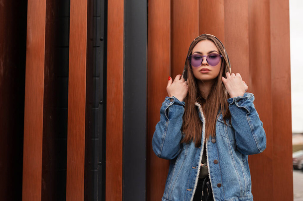 Moderne attraktive junge Frau in stylischer blauer Jeansjacke in militärischem Kapuzenpulli mit modischer lila Brille posiert neben einer metallenen Vintage-Wand im Freien. Trendiges Hipster-Mädchen in der Stadt. Straßenstil. - Foto, Bild