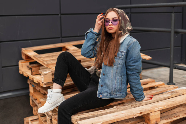 Urban cool giovane donna alla moda hipster in abiti casual alla moda giovani in occhiali viola alla moda posa in città. Modello di moda ragazza glamour si siede su pallet di legno per strada. Stile primaverile. - Foto, immagini
