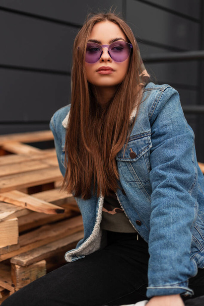 Modieus portret jonge vrouw hipster in vintage violette bril in stijlvolle blauwe vintage denim jas bij het grijze gebouw buiten. Mooi stedelijk meisjesmodel rust op de stad. Amerikaanse stijl. - Foto, afbeelding
