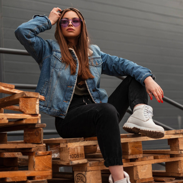Módní modelka mladá žena ve stylovém džínovém oblečení v módních fialových brýlích v kožených bílých botách odpočívá na dřevěných paletách u zdi ve městě. Pěkný trendy moderní dívka hipster venku. - Fotografie, Obrázek