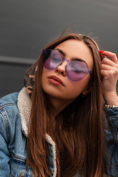 Stylisches Porträt einer jungen Frau in modischer lila Brille in blauer Jeansjacke in der Stadt. Attraktives Mädchen in lässiger Jugendkleidung sitzt mit geschlossenen Augen und genießt den frühlingshaften warmen Tag - Foto, Bild