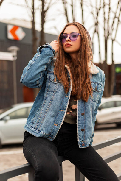 Стильна модель молодої хіпстерки в модних фіолетових окулярах в модній опуклій синій джинсовій куртці вирівнює довге волосся біля паркінгу на вулиці. Сучасний дівчина-хіпстер в старовинному одязі на місті
. - Фото, зображення