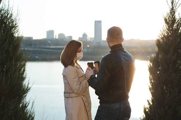 Young couple, a man and a woman in medical masks and gloves, drink coffee from disposable cups on the street and look at each other against the background of the river and the city. Quarantine - Foto, afbeelding