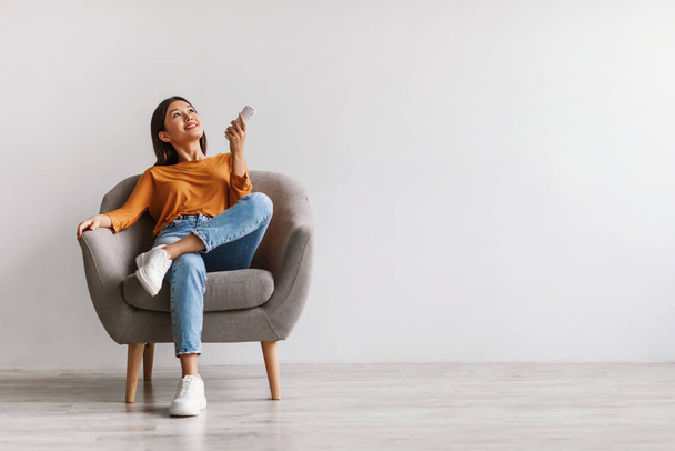 Happy young Asian woman with remote relaxing under air conditioner, sitting in armchair against white wall, mockup - Φωτογραφία, εικόνα