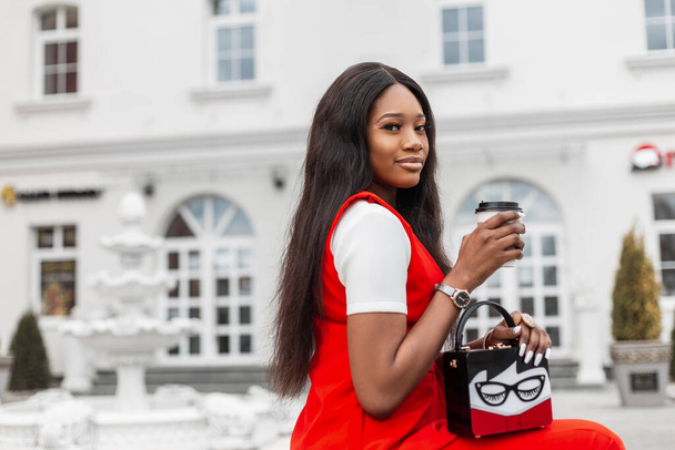 Modische schwarze junge Frau in modischen roten Kleidern mit Vintage-Handtasche mit Tasse Kaffee ruht auf Bank in der Nähe des weißen Gebäudes in der Stadt. Stilvolle hübsche Afrikanerin genießt Entspannung und Heißgetränk. - Foto, Bild