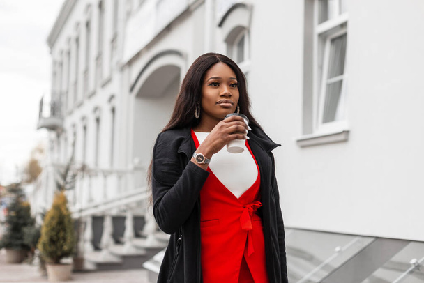 Prachtige jonge Afrikaanse vrouw met sexy lippen in rood-zwarte modieuze kleding loopt op straat en drinkt koffie. Mooi zwart meisje in stijlvolle outfit met kopje warme drank geniet lopen in de stad. - Foto, afbeelding