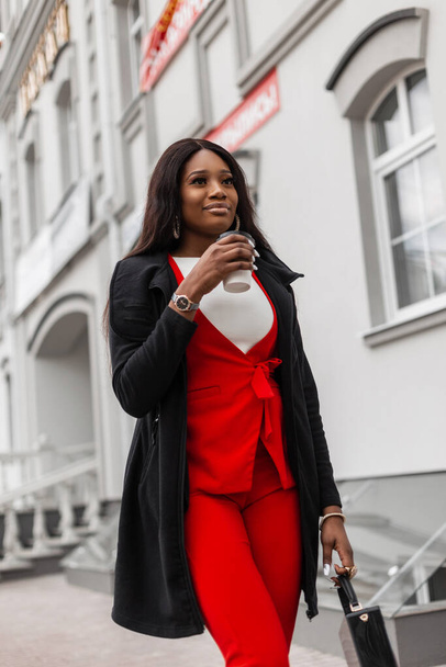 Μοντέρνο μοντέλο μοντέρνα νεαρή Αφρικανή γυναίκα σε κομψό κόκκινο κοστούμι στο σακάκι με φλιτζάνι καφέ βόλτες κοντά σε vintage κτίριο στην πόλη. Trendy χαριτωμένο ελκυστική μαύρο κορίτσι απολαμβάνει νόστιμο ποτό και τα πόδια. - Φωτογραφία, εικόνα