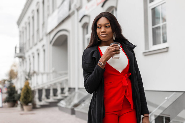 Moda mankeni oldukça hoş, şık kırmızı takım elbiseli, şık bir Afrikalı kadın şehrin klasik binasının yanında bir fincan kahveyle yürüyor. Modaya uygun seksi siyah kız lezzetli içeceklerden ve dışarı çıkmaktan hoşlanıyor.. - Fotoğraf, Görsel