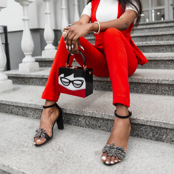 Elegante estilo de ropa femenina de moda. Mujer africana negra delgada en ropa de abrigo de negocios roja con bolso de moda y zapatos de verano se sienta en los escalones - Foto, imagen