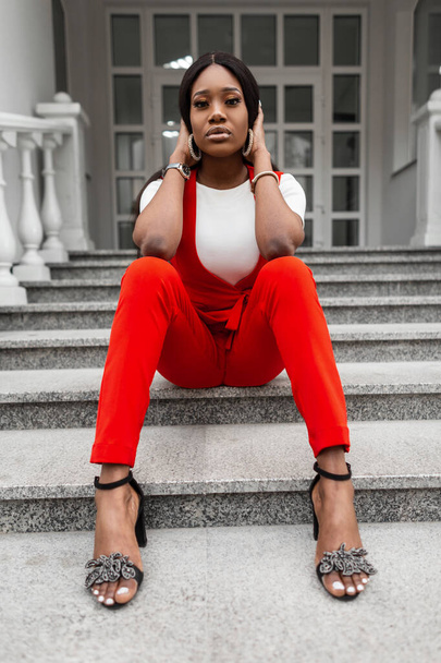 Stilvolle junge schwarze Frau mit sexy Lippen in modisch eleganter Kleidung in stylischen Sandalen posiert an einem Sommertag auf einer steinernen Vintage-Treppe in der Stadt. Attraktives Mode-Modell schwarzes Mädchen im roten Anzug. - Foto, Bild