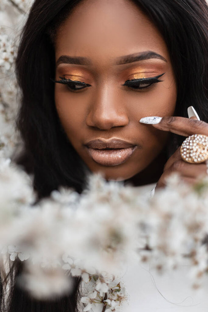 Nahaufnahme Porträt einer jungen schönen schwarzen Frau mit sauberer Haut und Maniküre in der Nähe von Blumen auf der Straße. Gesunder und schöner Lebensstil - Foto, Bild