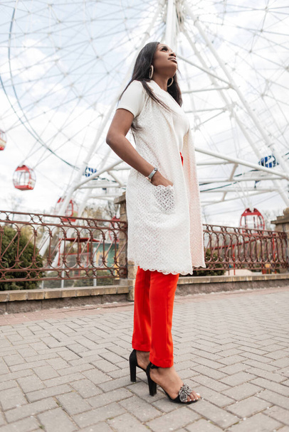 Fashionable black young woman in long white vest in red vintage trousers in summer sandals stands in city on the background of Ferris wheel. Attractive stylish african girl walks on street. Fashion. - Photo, Image