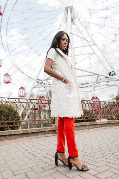 Trendy young African woman in stylish red-white summer clothes in shoes stands on street near amusement park. Black girl in elegant outfit rests on backdrop of vintage ferris wheel on bright day. - Photo, Image