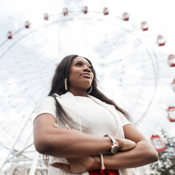 Trendy young african woman with sexy lips in white fashionable clothes poses in park near attractions. Fine urban African woman in elegant outfit on background of ferris wheel and sky in bright day. - Photo, Image