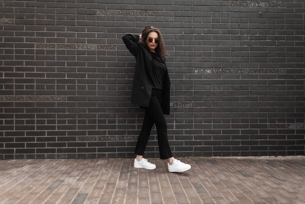Μοντέλα μόδας νεαρή γυναίκα με μοντέρνα γυαλιά ηλίου σε κομψά casual παιδικά ρούχα σε λευκά sneakers που ποζάρουν κοντά σε vintage μαύρο τούβλο τοίχο στο δρόμο. Ευρωπαϊκή αστική κοπέλα με μαύρη στολή σε εξωτερικούς χώρους. Στυλ. - Φωτογραφία, εικόνα
